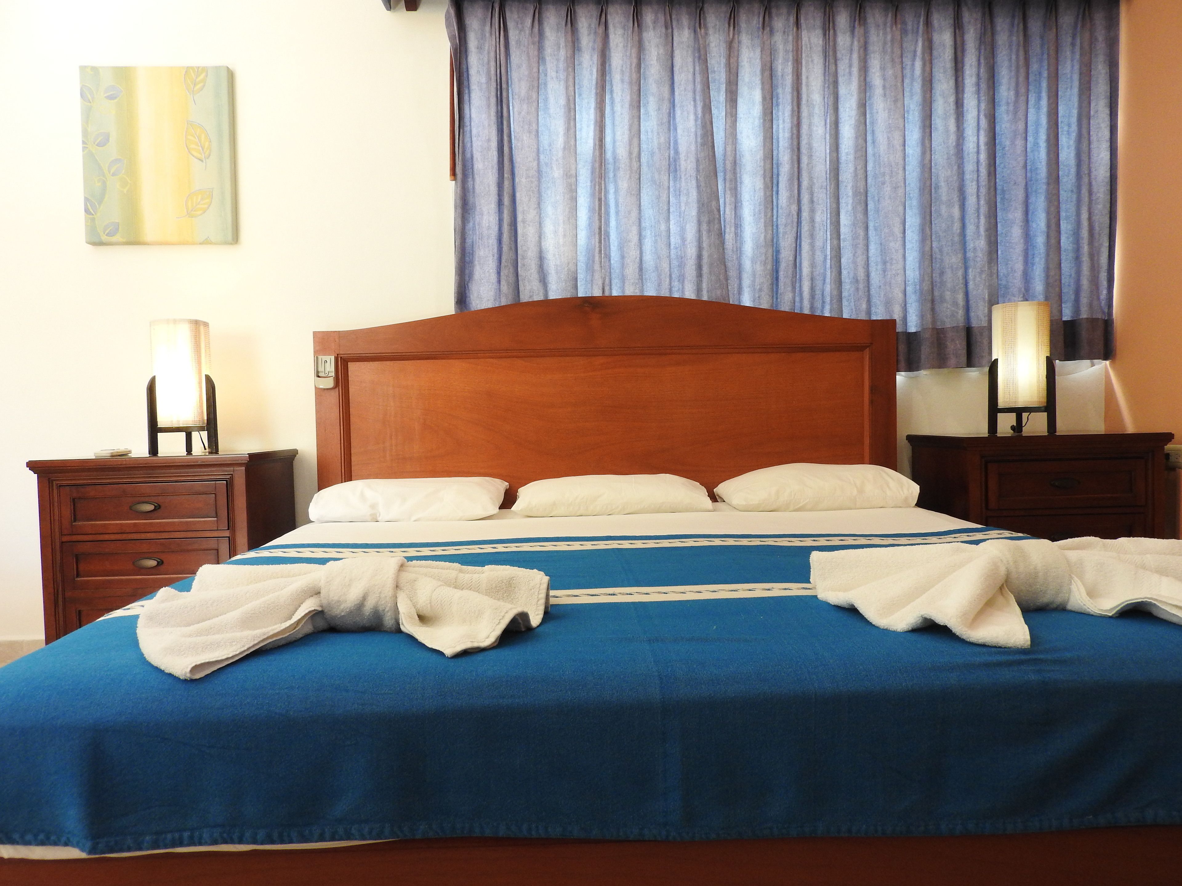 A neatly made bed with a blue bedspread and white pillows, two bedside tables with lamps, and a painting on the wall.
