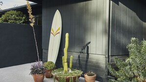 Een modern gebouw met een donkere gevel, een surfplank ertegen en diverse potplanten ervoor.