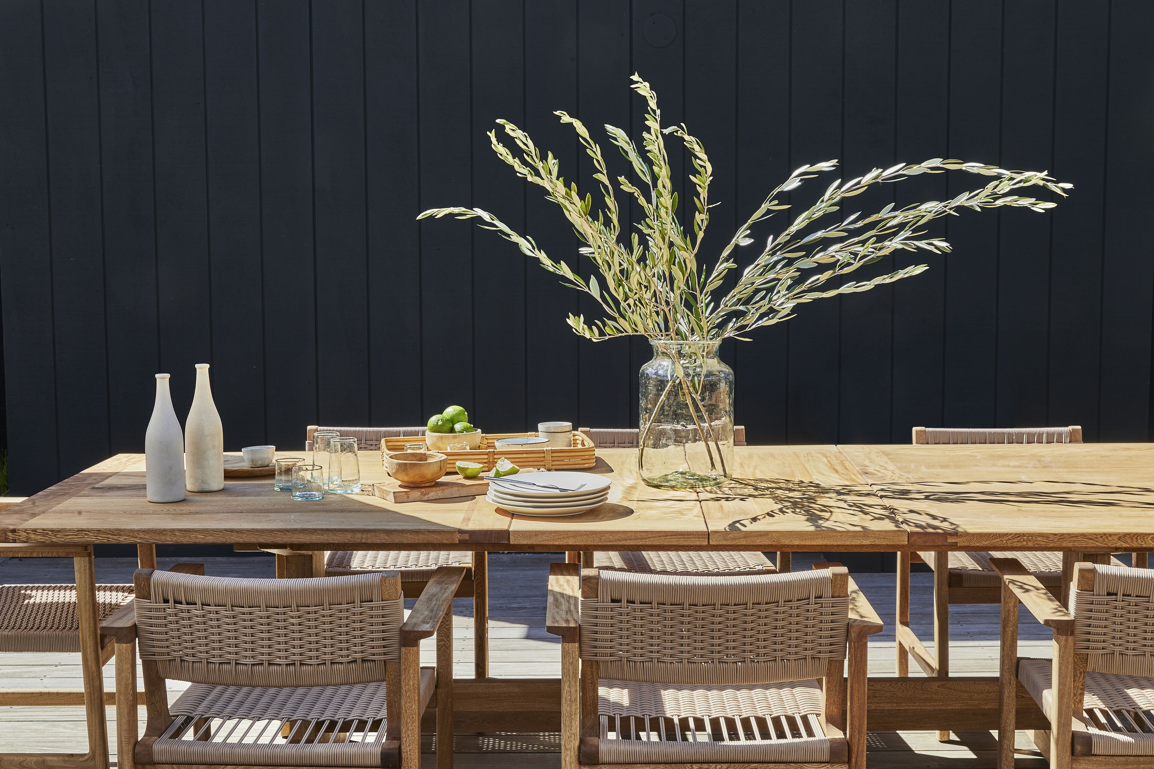 A wooden dining table set with wicker chairs, a vase with dried plants, and a glass of water.