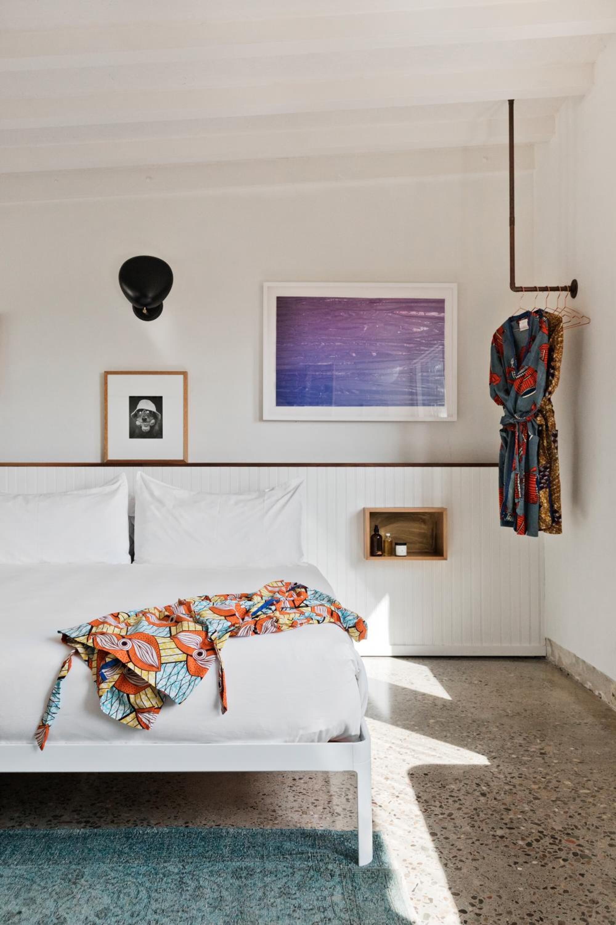 Un dormitorio con una cama, un cuadro enmarcado, una bata colgada y una lámpara fijada a la pared.