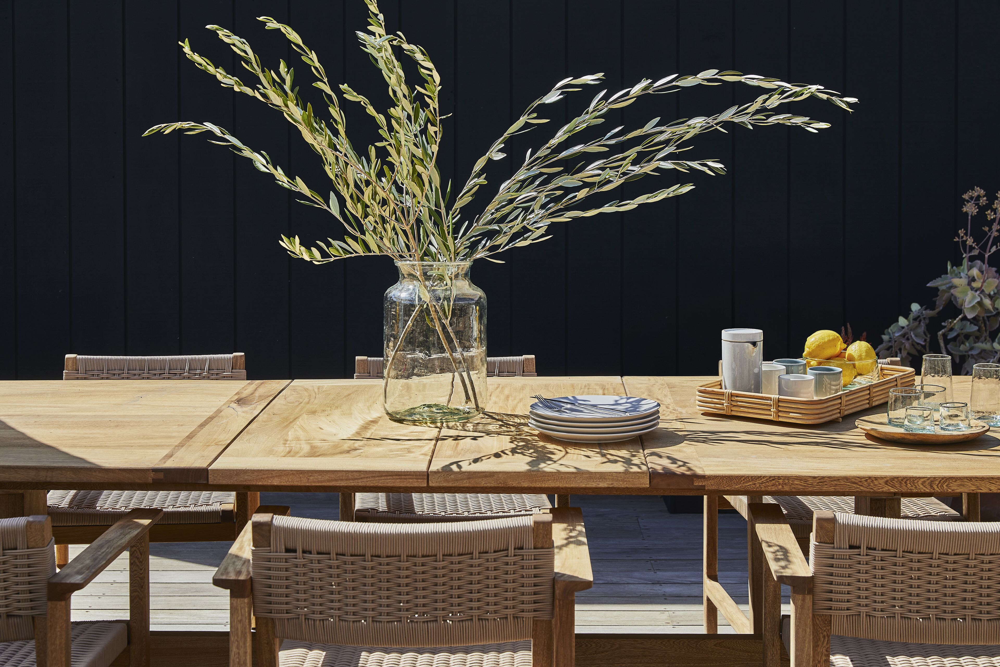 Una mesa de comedor de madera con sillas de mimbre, un florero de vidrio transparente con plantas secas y una cesta de frutas con limones.