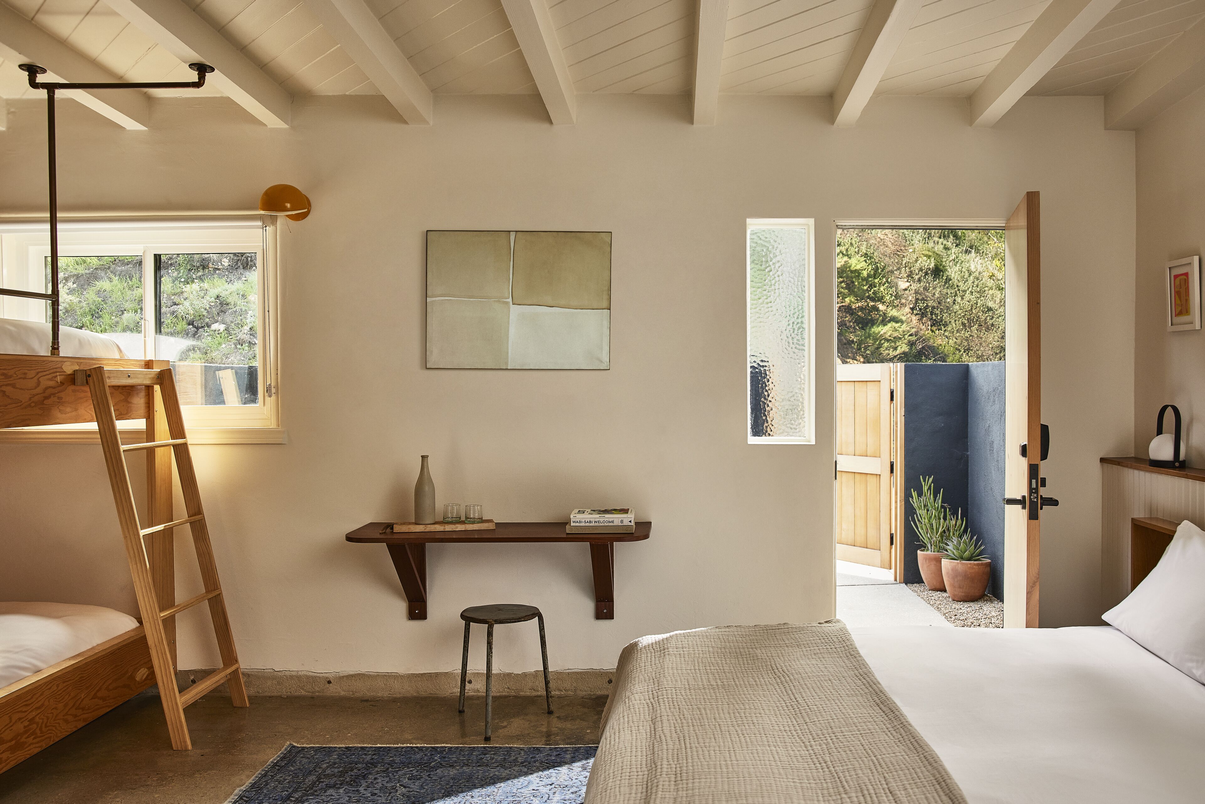 A bedroom with a bunk bed, a desk with a chair, a small stool, a bedside table with a vase, and a window with a view of greenery.