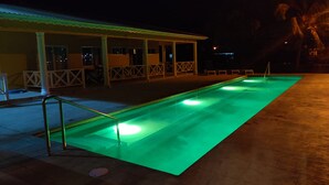 Una piscina rectangular con un borde iluminado en verde, una zona de estar techada con barandillas, y un edificio con tejado de baldosas al fondo.