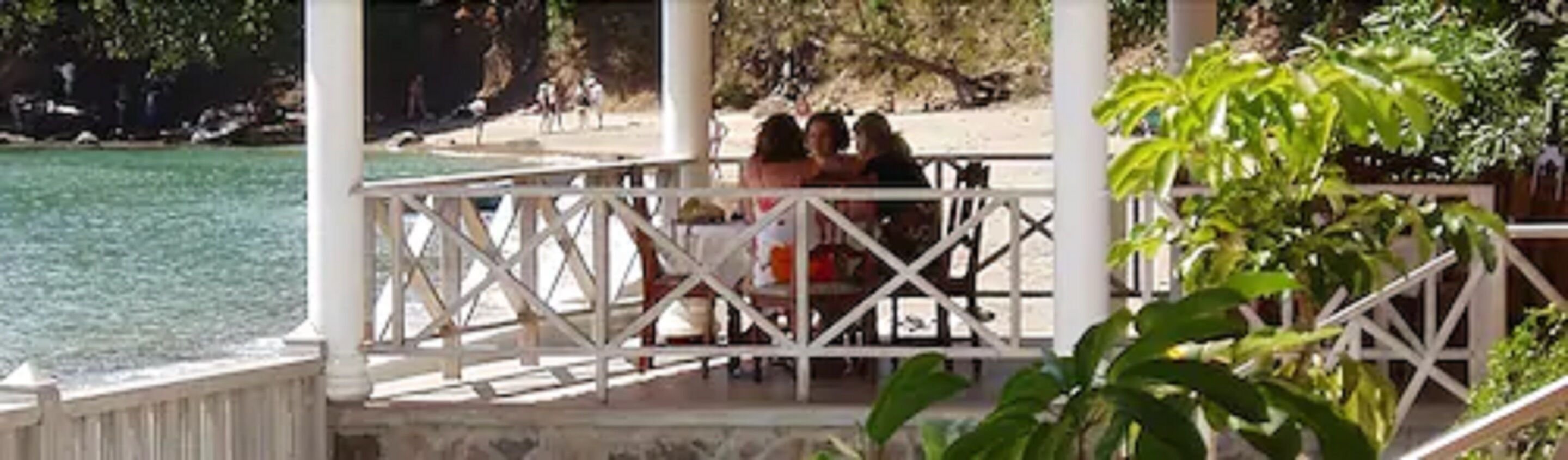 A covered outdoor area with a view of the sea, a white railing, and some greenery.