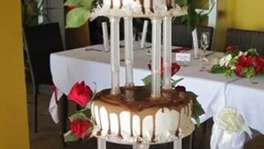Un pastel de tres niveles con glaseado de chocolate y rosas rojas sobre un mantel blanco.