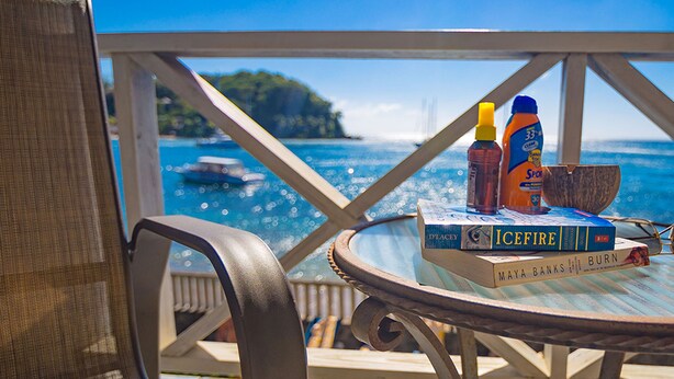 Una mesa con libros y botellas de protector solar en un balcón con vista al agua.