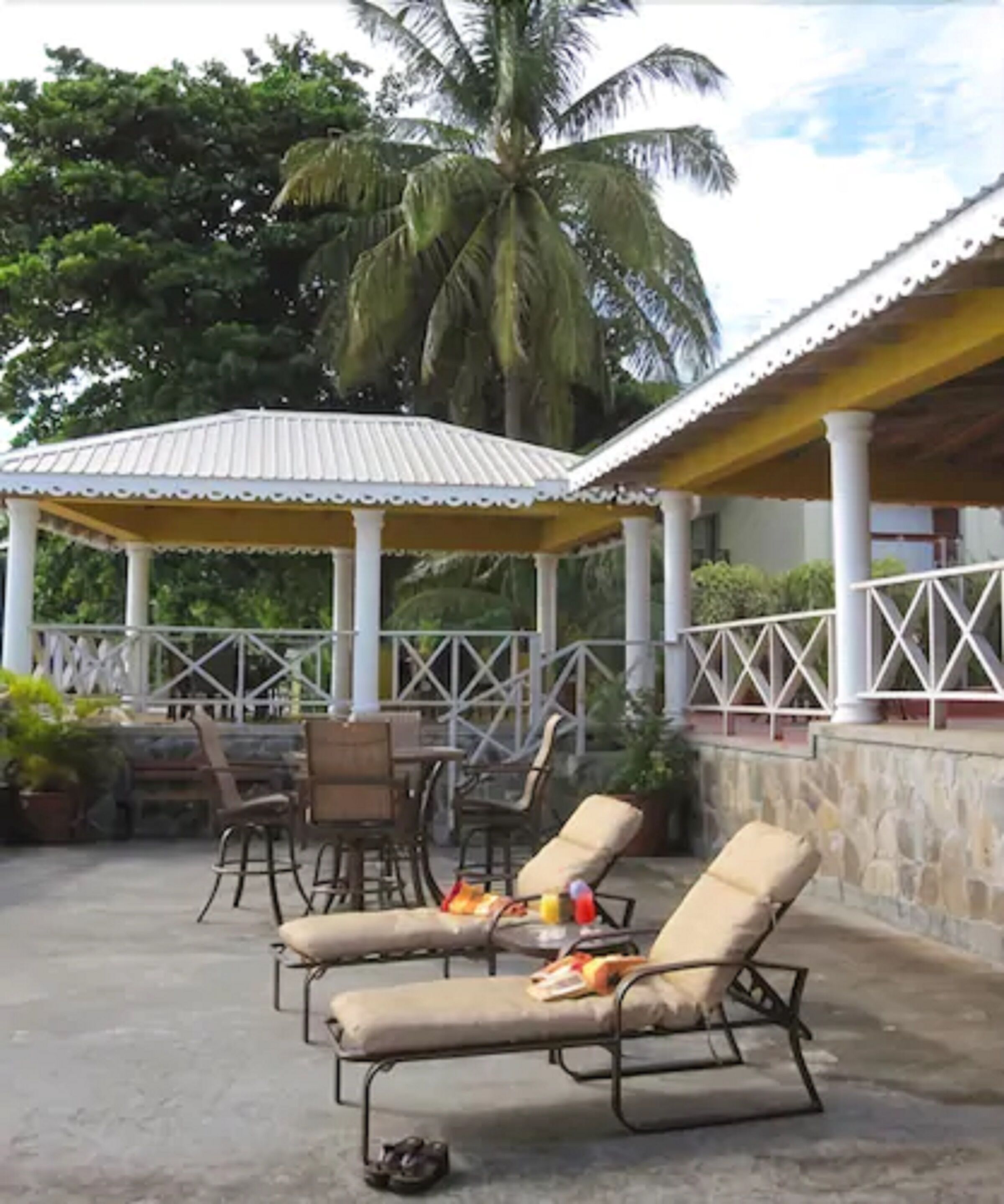 A covered outdoor seating area with lounge chairs and a table, surrounded by a stone wall and palm trees.