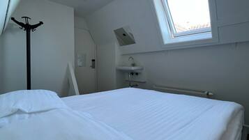 Een slaapkamer met een bed, een raam, een radiator, een kapstok en een klein bureau met een spiegel.