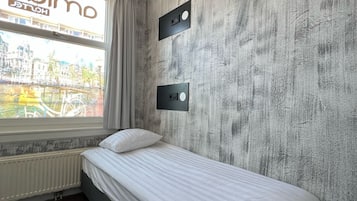 A hotel room with a bed, a window with a view of a cityscape, a radiator, and a ceiling fan.