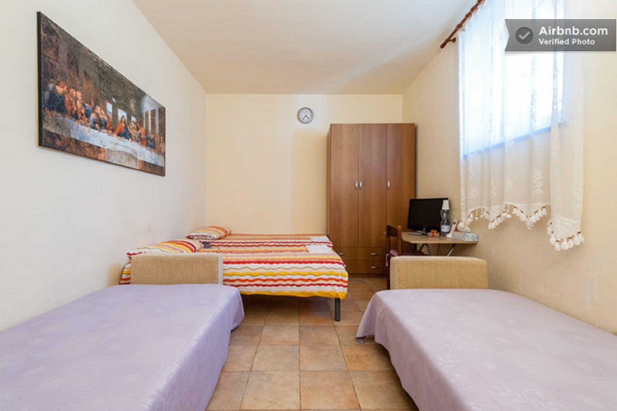 A small, cozy room with two single beds, a wooden wardrobe, a desk with a chair, and a painting on the wall.