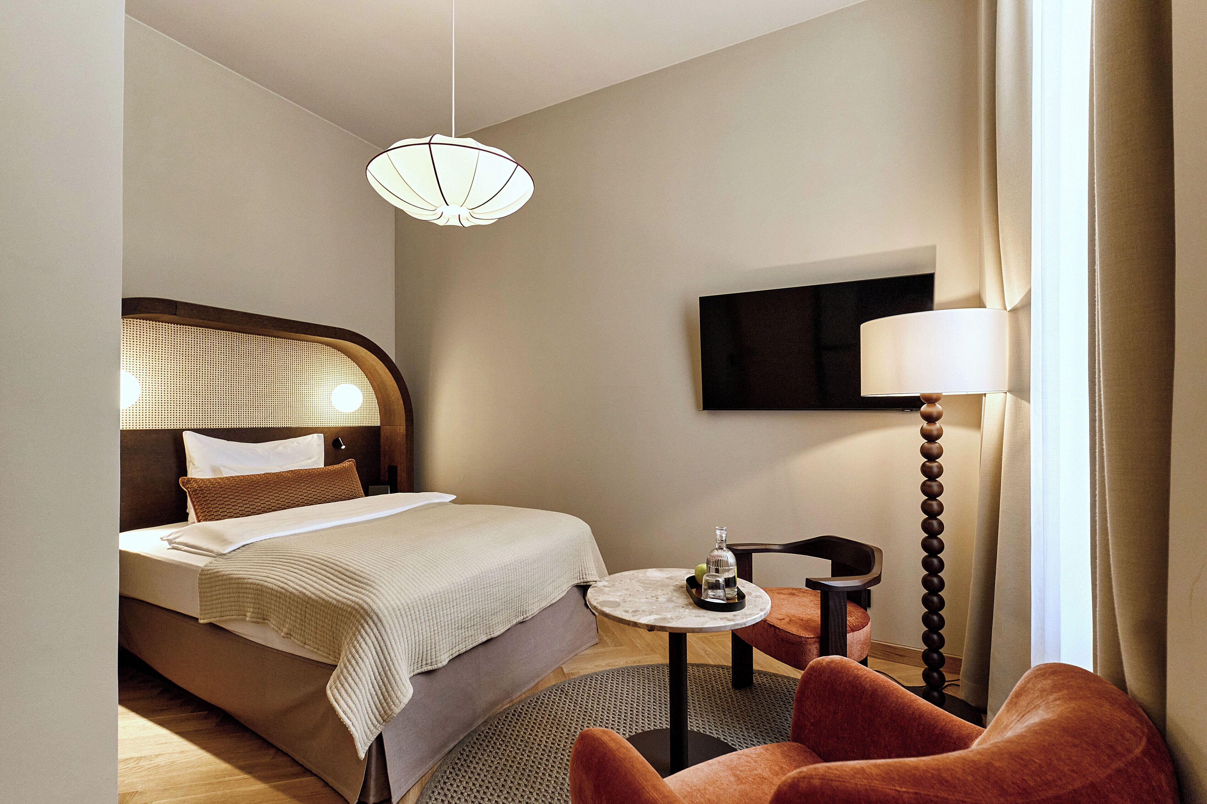 A hotel room with a bed, bedside table, lamp, chair, and a television on the wall.