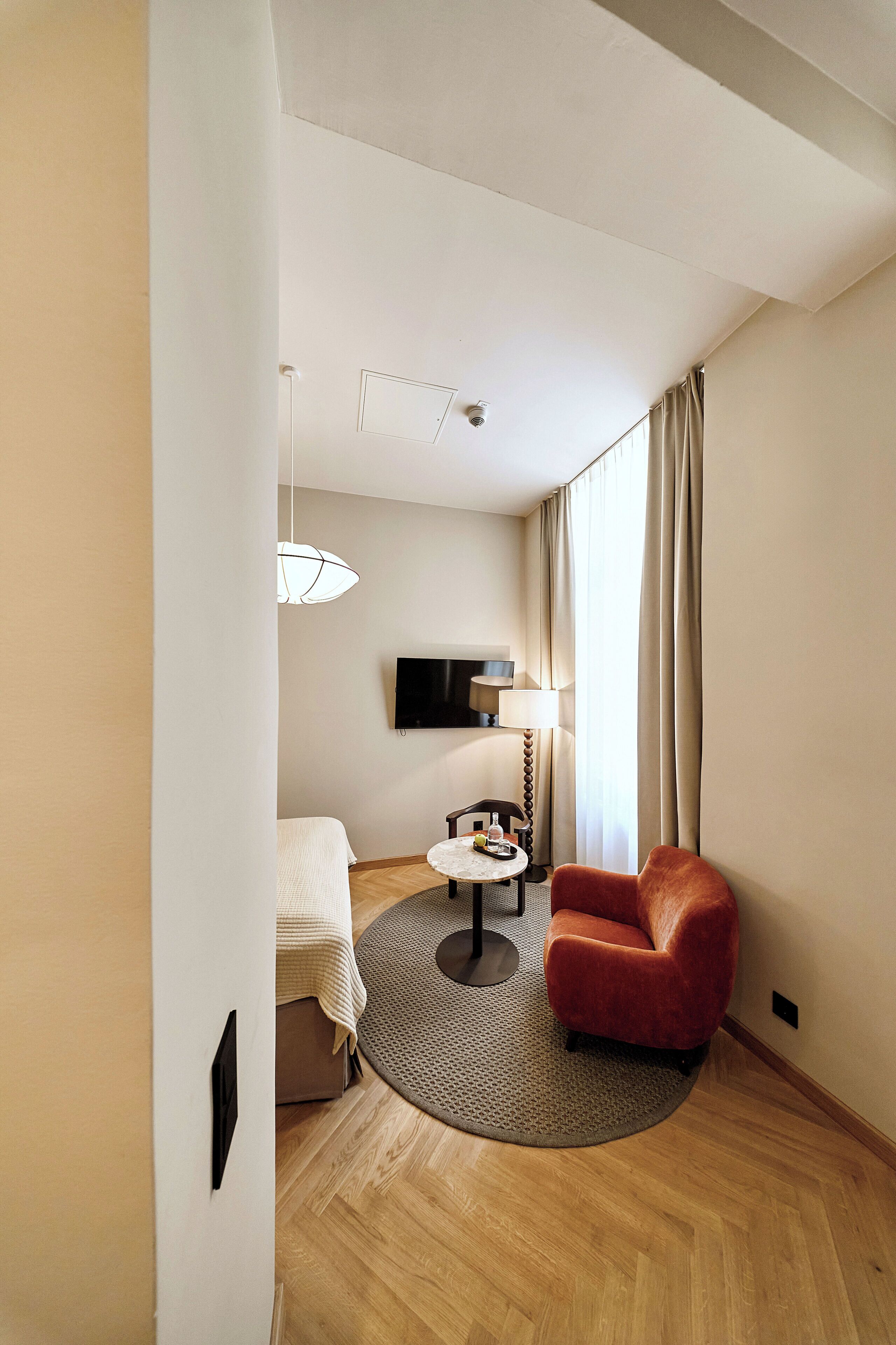 A hotel room with a bed, a round table with a lamp, a red chair, a television on the wall, and a large window with curtains.