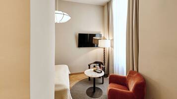 Una habitación de hotel con una cama, una mesa redonda con lámpara, una silla roja, un televisor en la pared y un ventanal con cortinas.