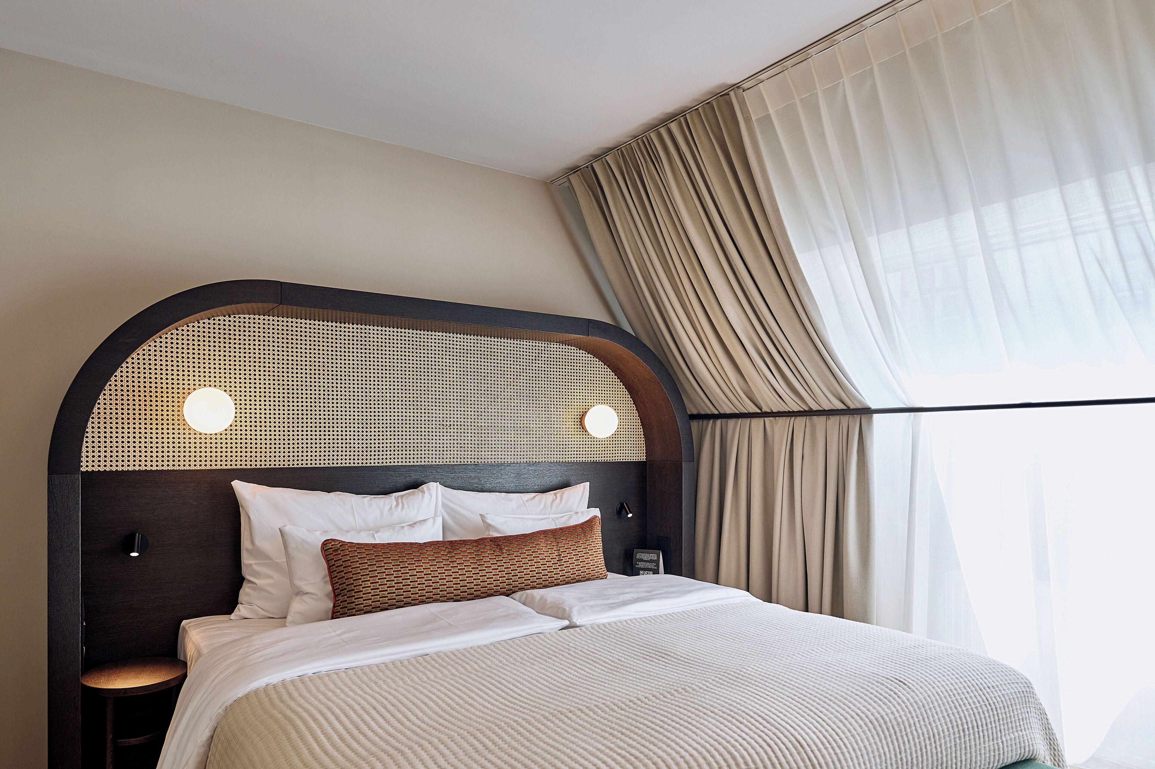 A hotel room with a large bed, a headboard with a woven design, two bedside lamps, and a view of the outside through sheer curtains.