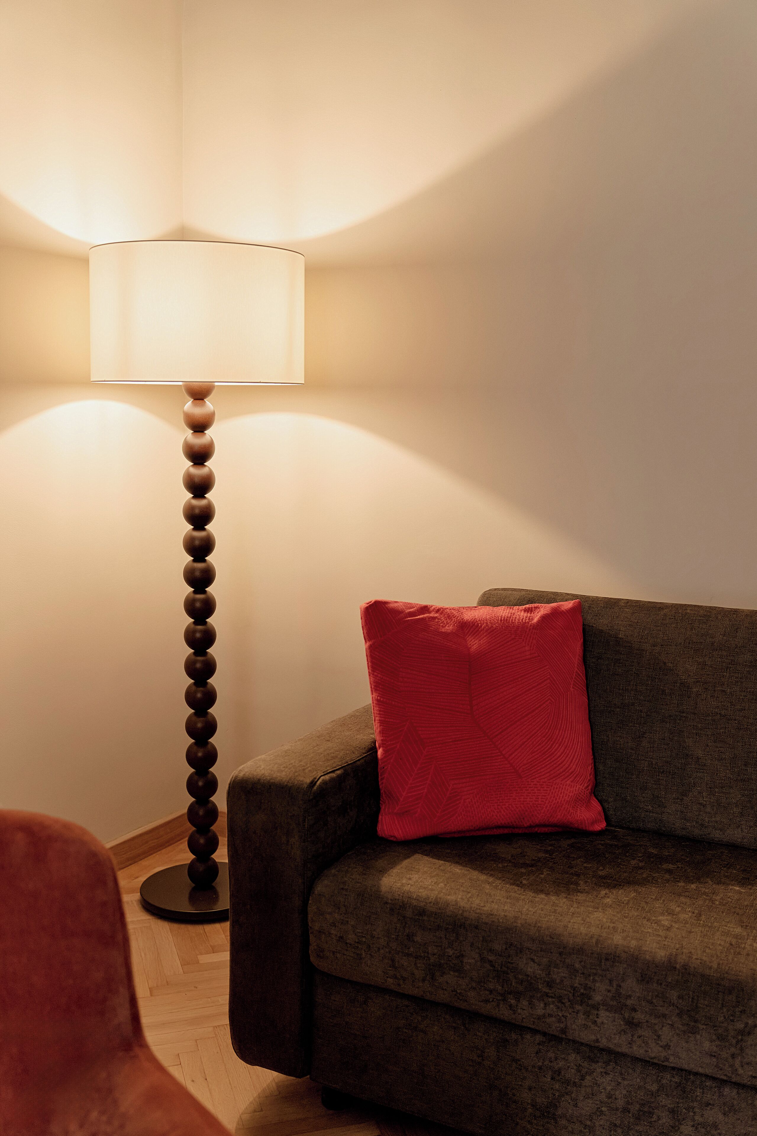 A room with a floor lamp, a sofa with a red pillow, and a wooden floor.