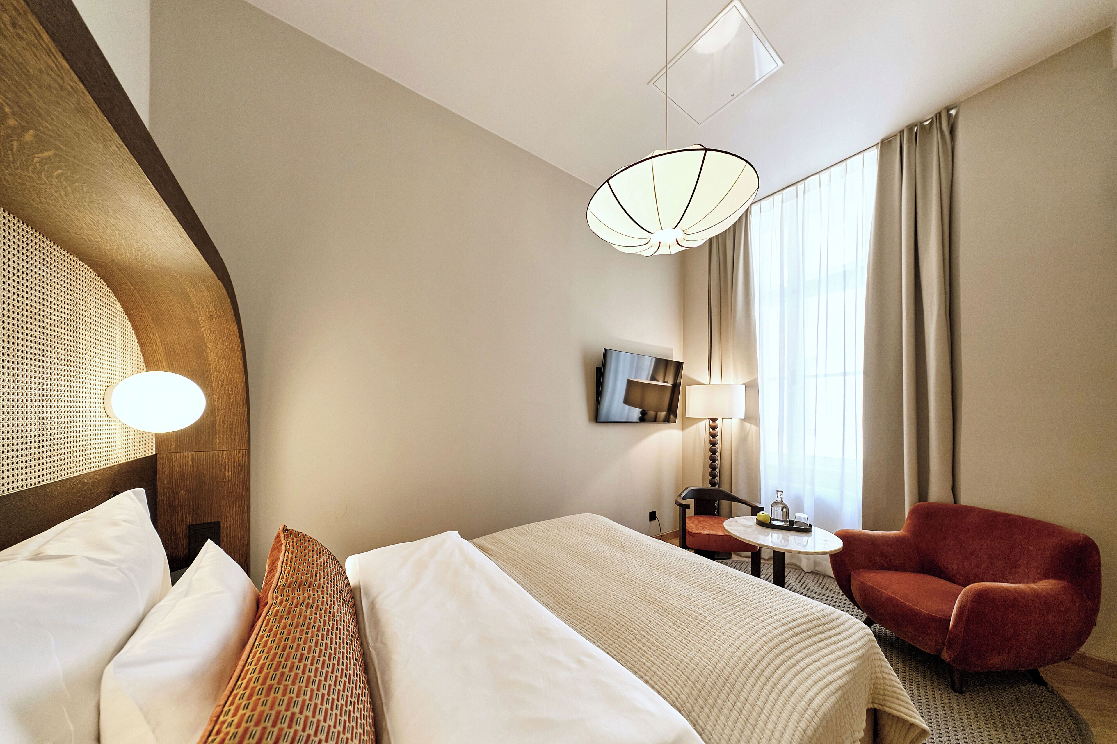 A hotel room with a bed, a red armchair, a small table with a lamp, and a large window with curtains.