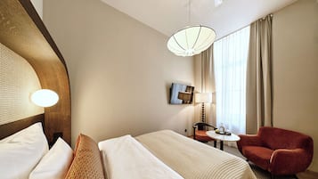 Una camera d'albergo con un letto, una poltrona rossa, un tavolino con una lampada e un'ampia finestra con tende.