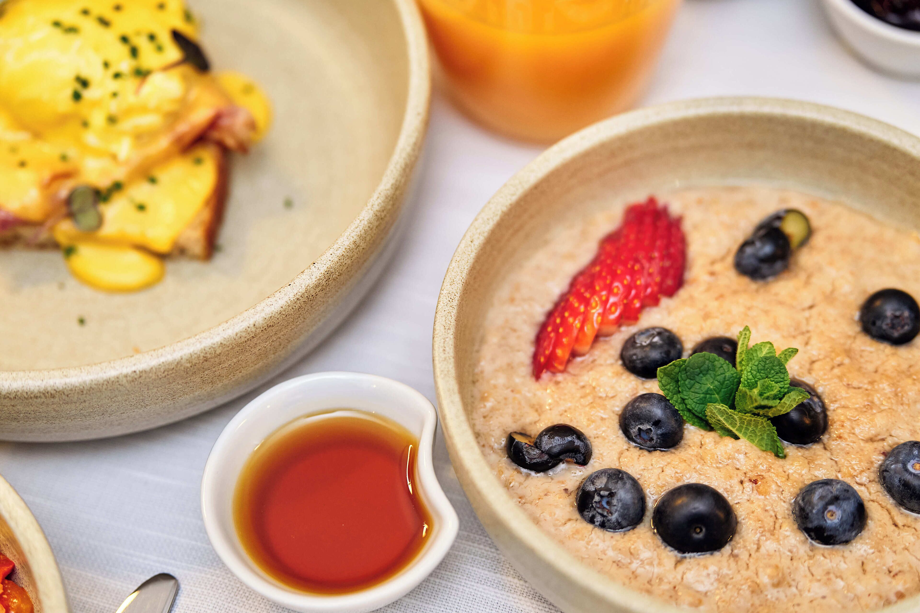 Une table dressée pour le petit-déjeuner, avec un bol de porridge garni de myrtilles, d’une fraise et de quelques feuilles de menthe, accompagné d’un petit ramequin de sirop.