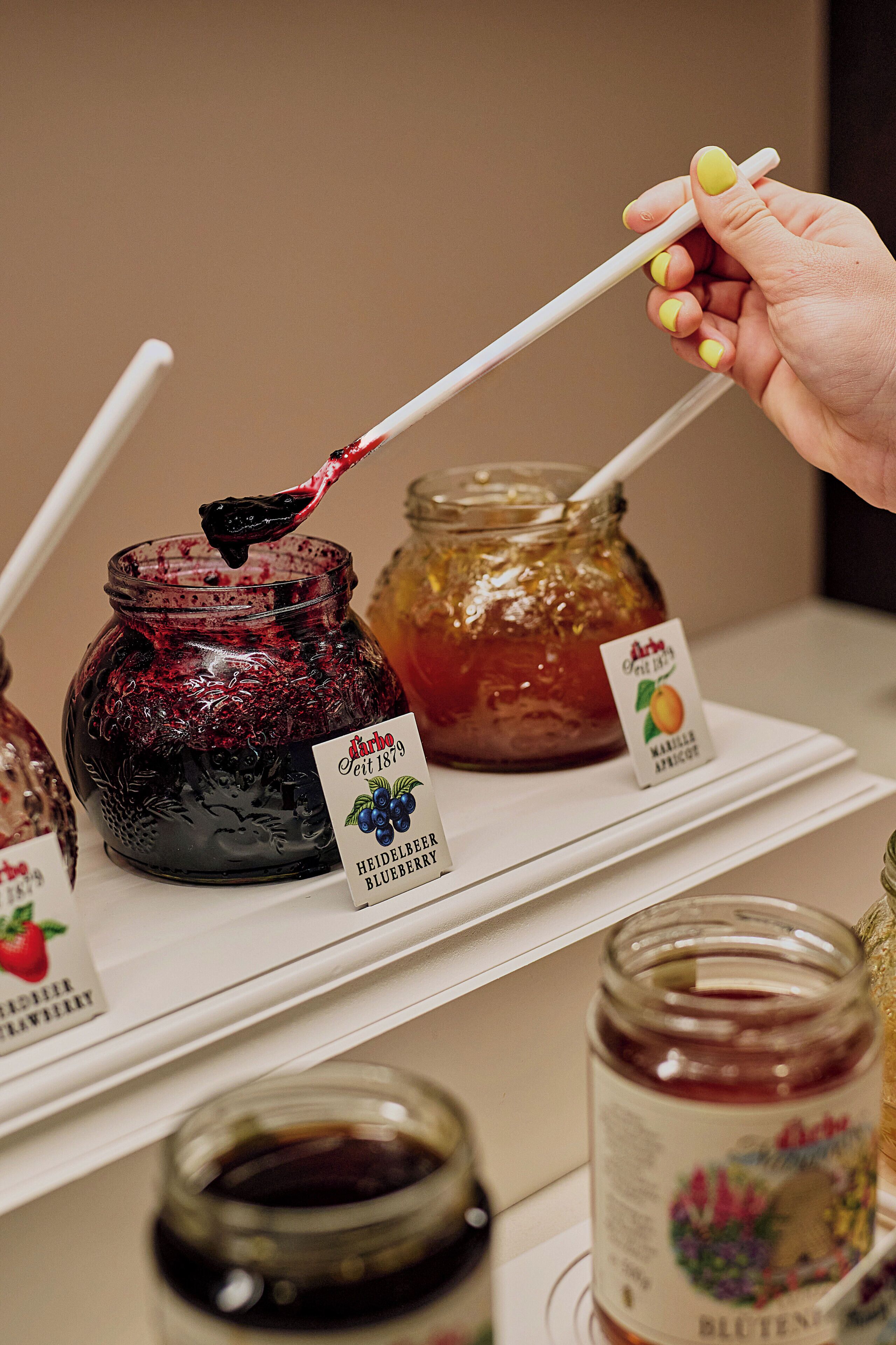 A hand holding a chopstick with a piece of food, a jar of blueberry jam, and a jar of apricot jam on a shelf.
