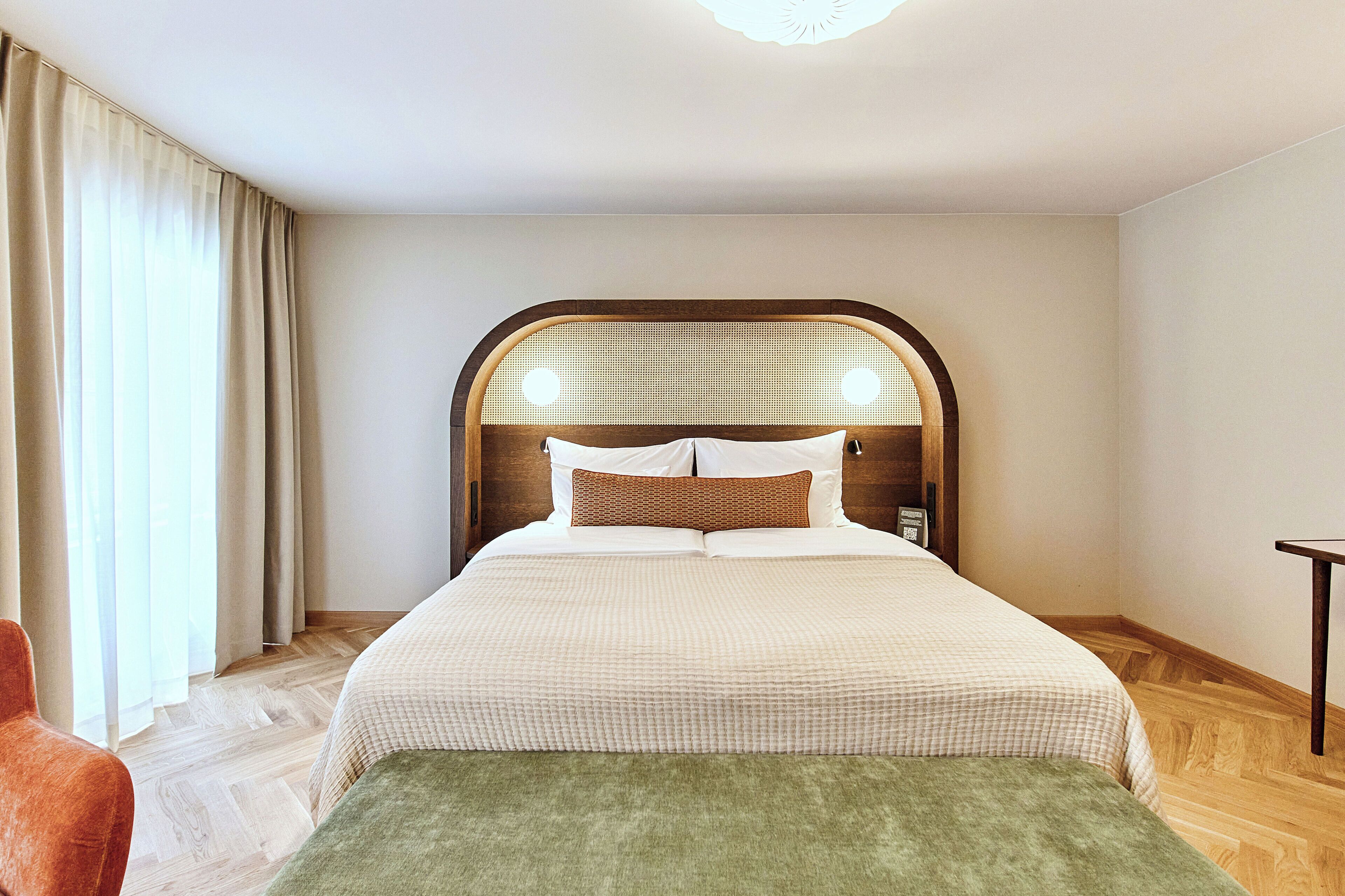 A hotel room with a large bed, a wooden headboard, a small table, and a window with curtains.