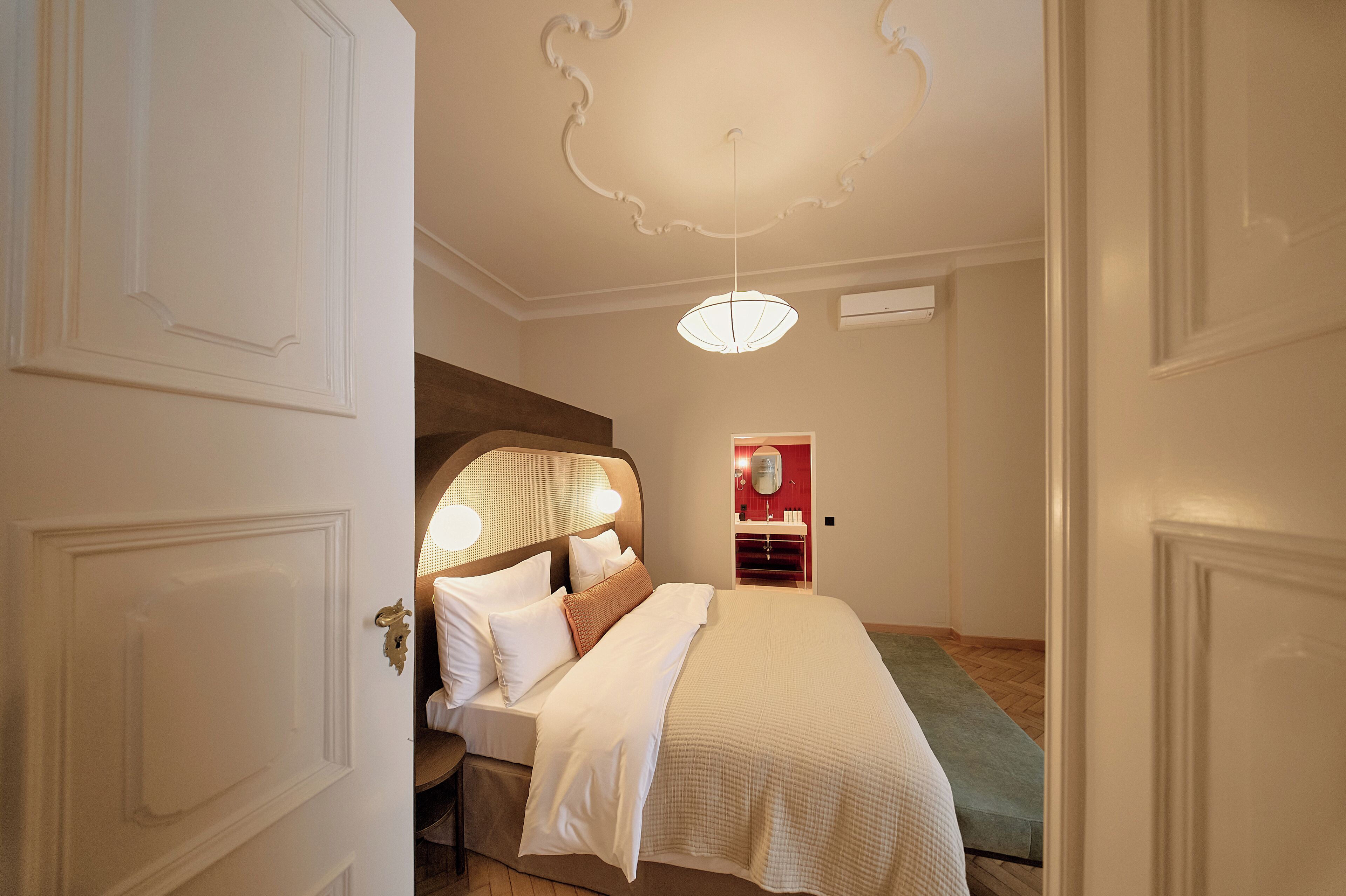 A hotel room with a bed, a nightstand, a mirror, a red wall, and a ceiling light.