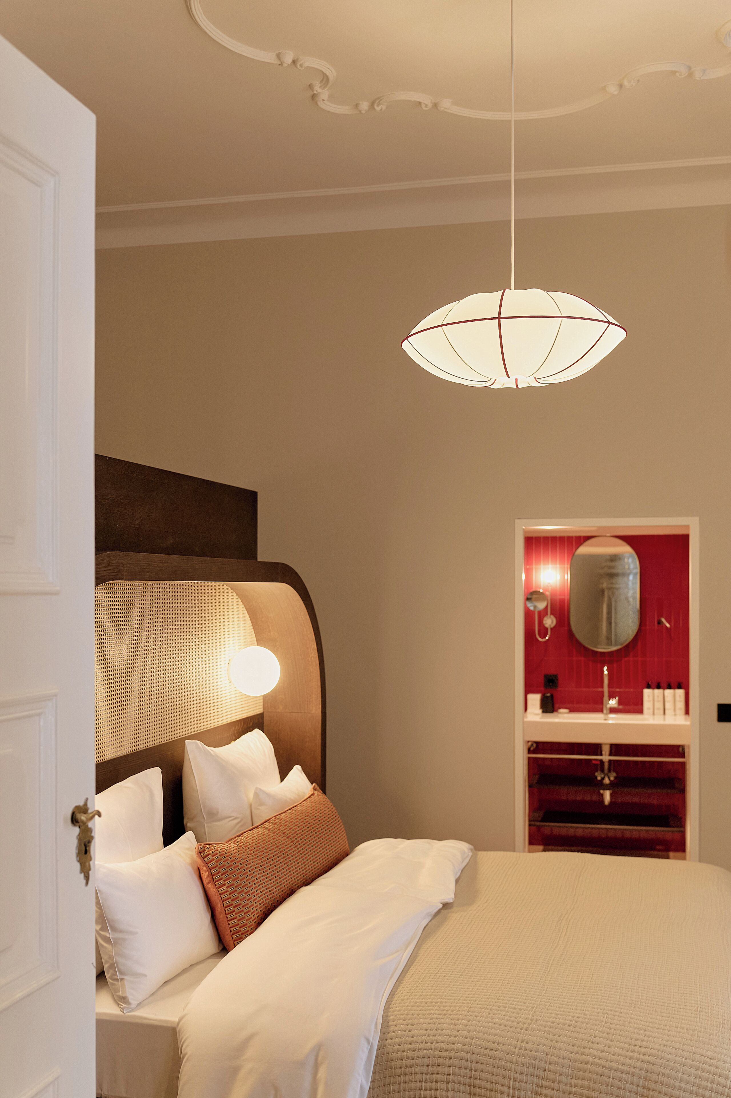 A neatly made bed with pillows, a bedside table, a hanging light fixture, and a red-framed mirror above a sink.