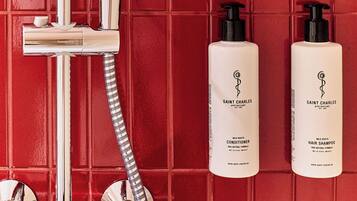 Baño con pared de azulejos rojos, cabezal de ducha cromado y dos botellas de champú y acondicionador Saint Charles.
