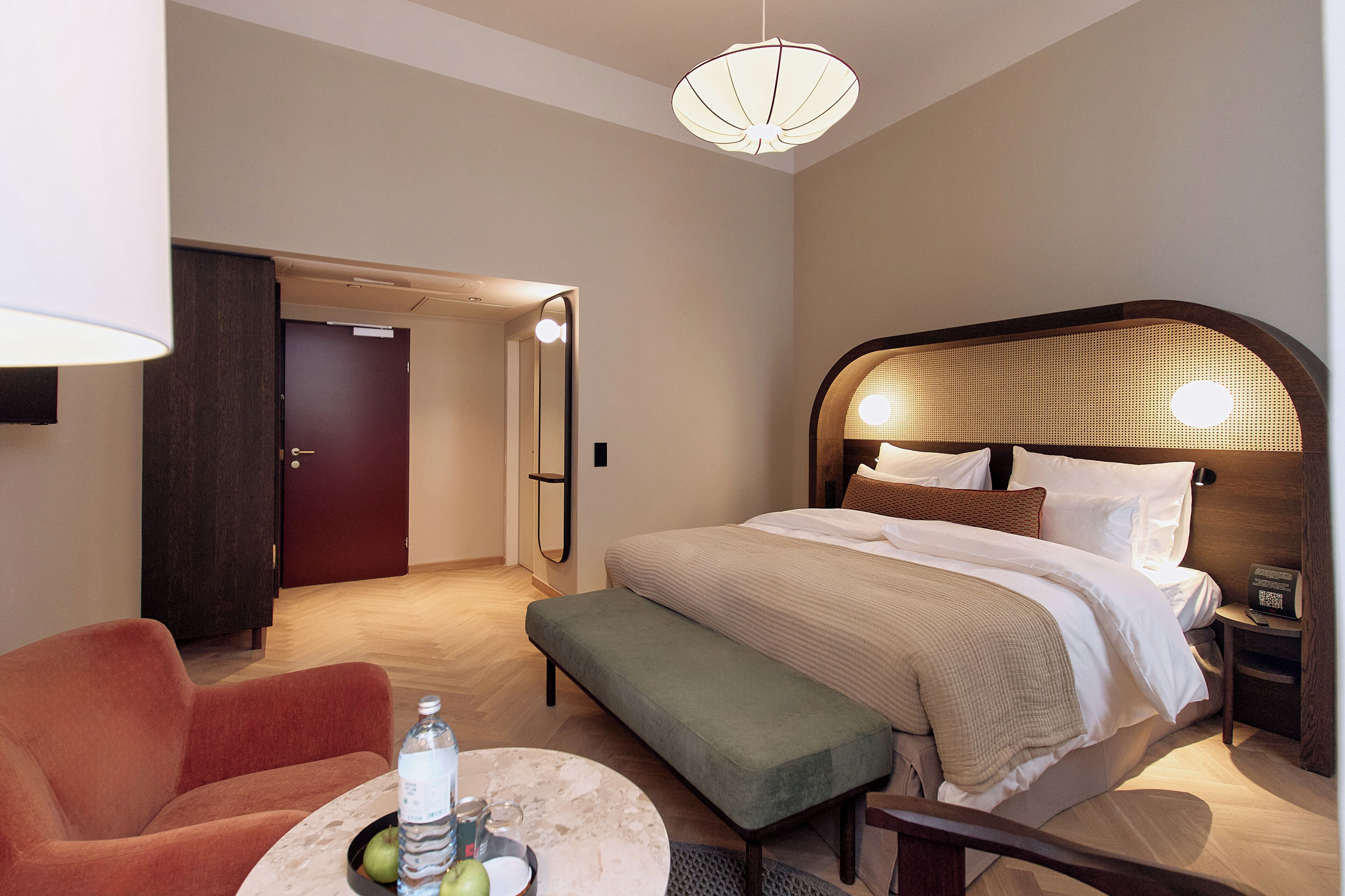 A hotel room with a bed, bedside table, lamp, and a seating area with a chair and bench.