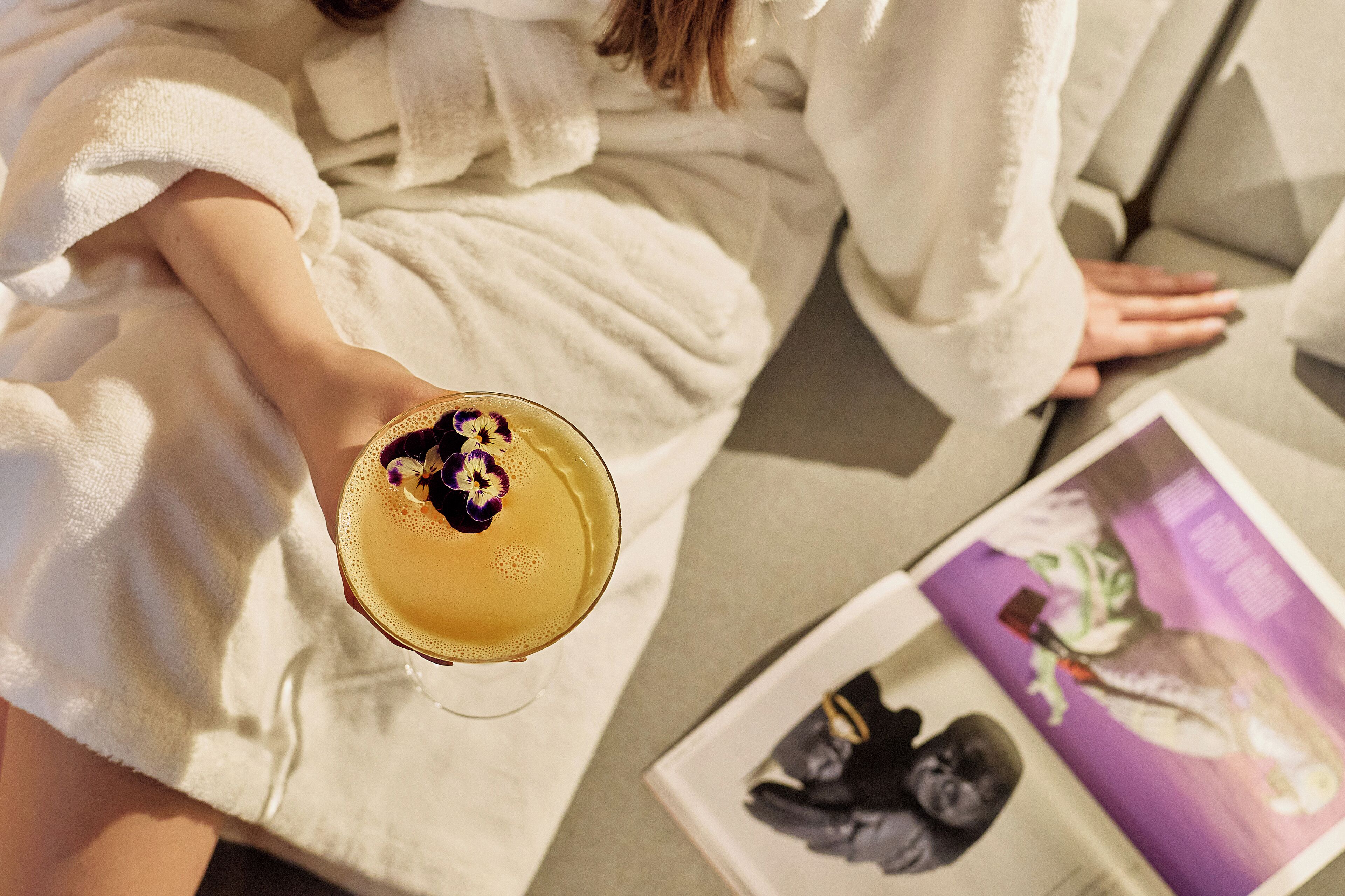 A person in a white bathrobe holding a drink with a flower garnish, sitting next to a magazine with a purple cover.