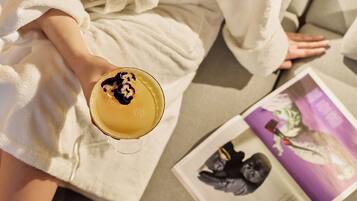 A person in a white bathrobe holding a drink with a flower garnish, sitting next to a magazine with a purple cover.
