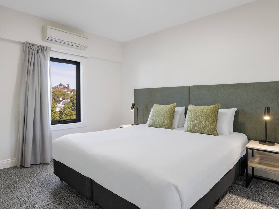 Quarto de hotel com uma cama grande, mesa de cabeceira com luminária, janela com cortinas e aparelho de ar condicionado.
