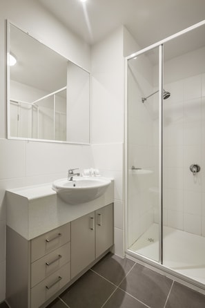 Features a wall-mounted mirror, sink, and drawers, along with a glass-enclosed shower area.