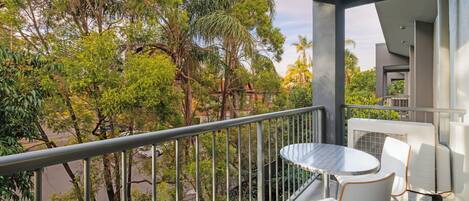 A balcony with a round table and two white chairs, overlooking a street with trees and a parked car.