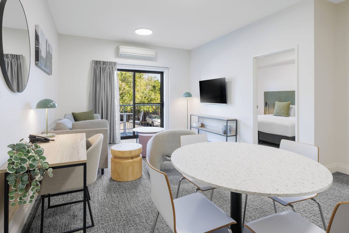 A modern hotel room with a round table, a sofa, a TV, and a balcony with a view of trees.