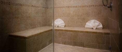 A shower area with beige tiles, a bench, and two towels on a shelf.