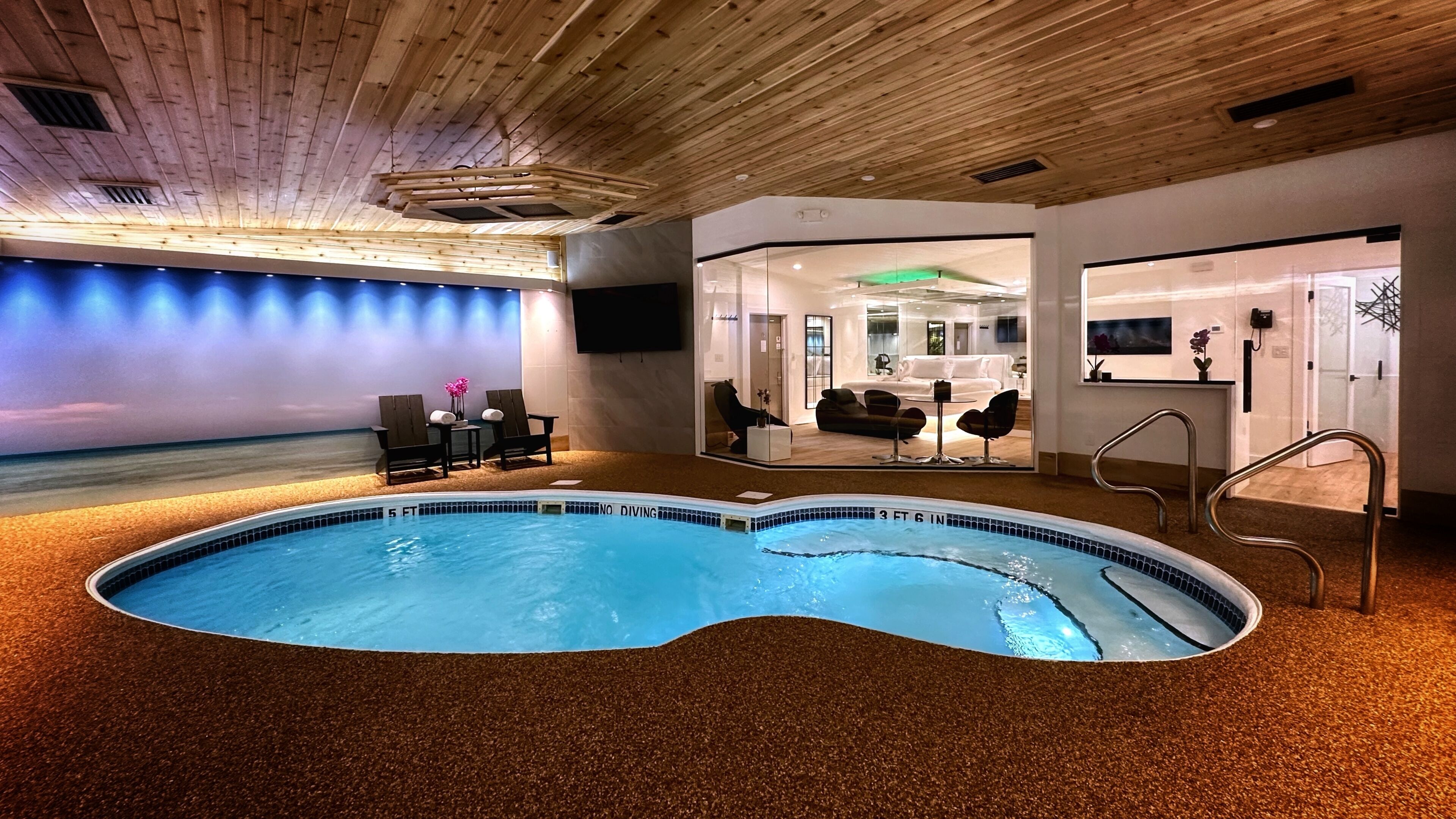 An indoor swimming pool with a wooden ceiling, a lounge area with chairs, and a view of a room with a bed and a sofa.