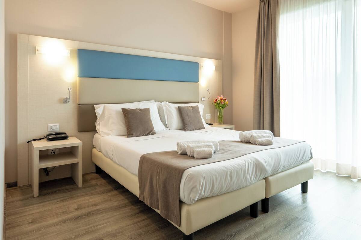 A hotel room with a bed, bedside tables, a nightstand, a wall-mounted light, and a window with curtains.