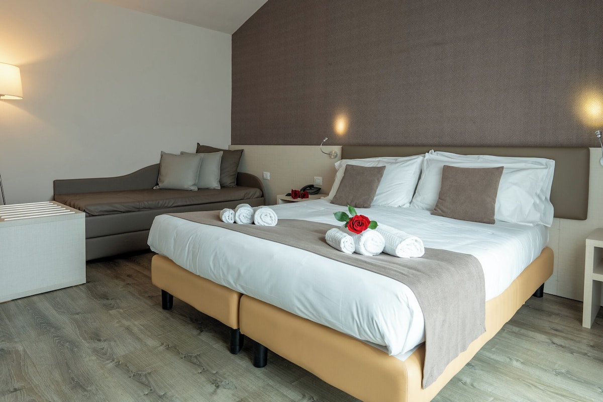 A modern hotel room with a bed, a sofa, a bedside table, and a lamp.
