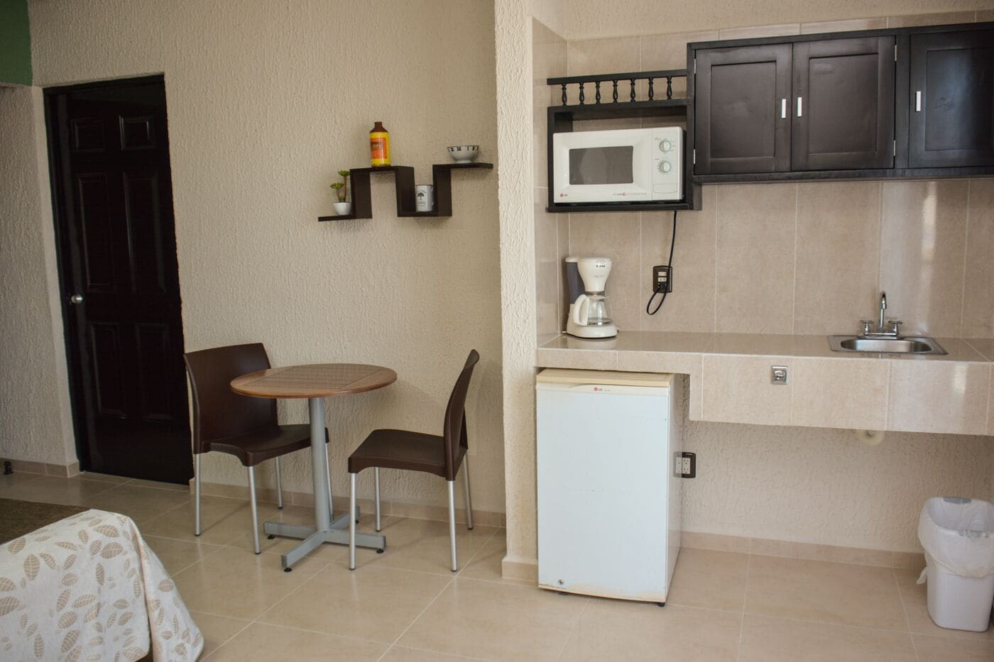 Cocina compacta equipada con microondas, cafetera y un refrigerador pequeño. Hay una mesa de comedor con sillas y una puerta negra al fondo.