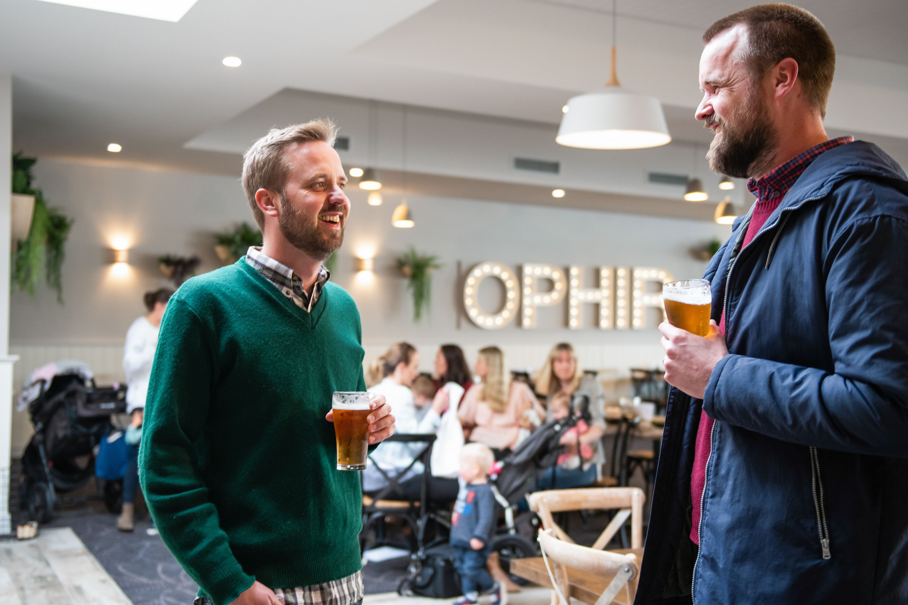 Two men holding glasses of beer in a social setting with other people in the background.