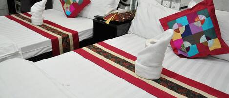 A hotel room with three beds, each with a white comforter and a red and black striped bed runner. There are towels placed on each bed, and a colorful quilted pillow on one of the beds.