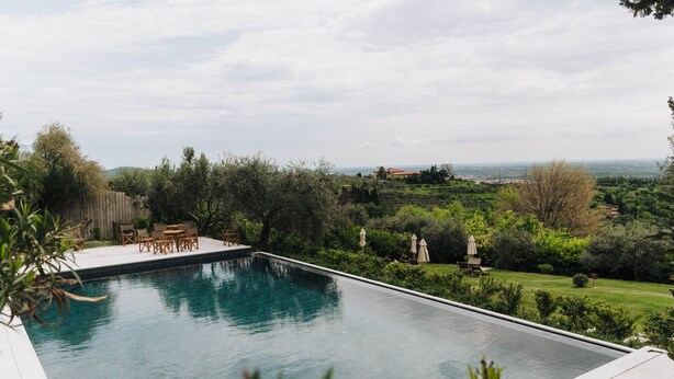 A rectangular swimming pool with a wooden deck and outdoor furniture, surrounded by lush greenery and a view of distant hills. - Corte San Mattia - Agriturismo in Verona (Verona)