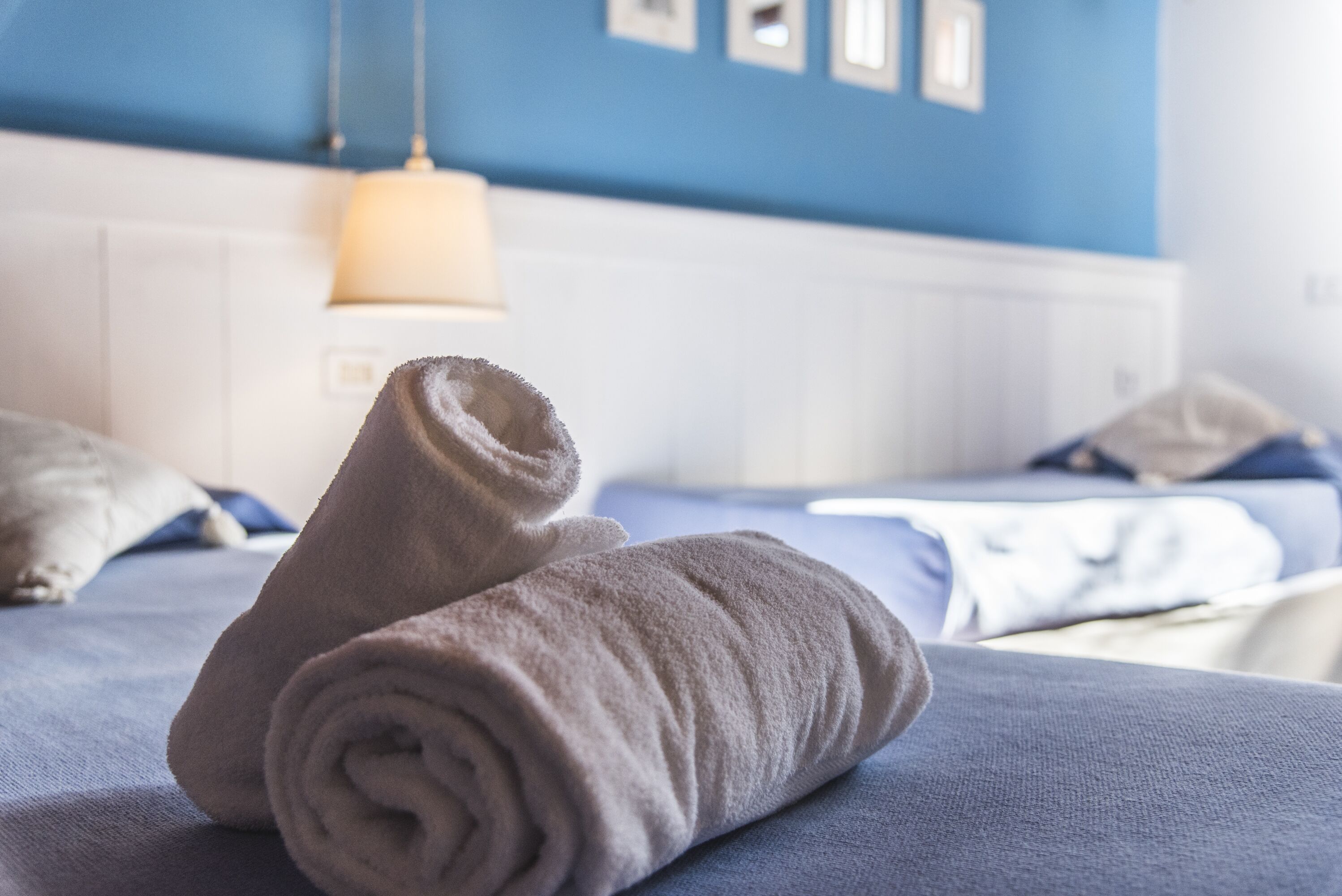 A neatly made bed with a rolled towel in the foreground, a blue wall, and a hanging light fixture in the background.