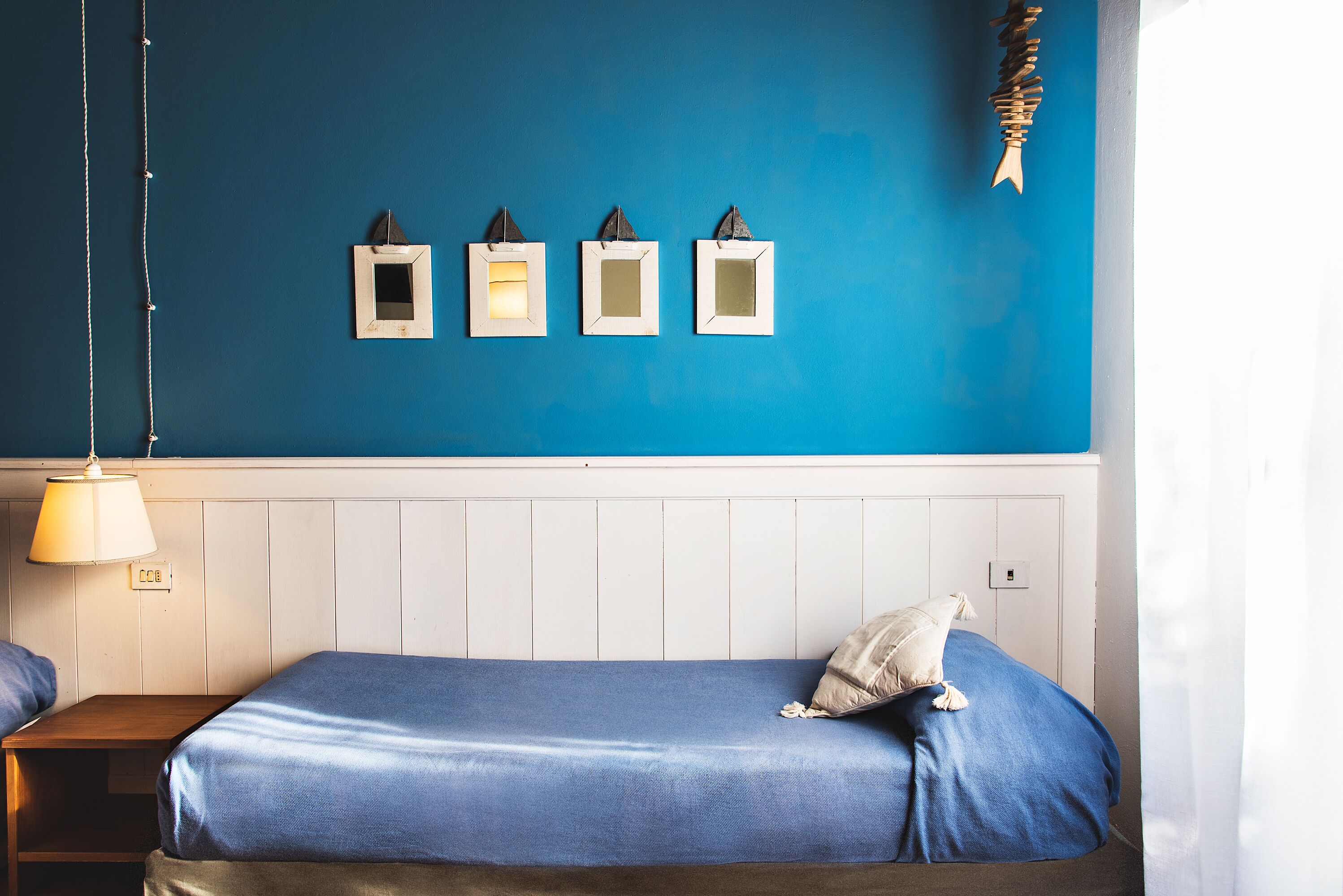 Une pièce avec un mur bleu, un lit recouvert d’une couverture bleue, une table de chevet avec une lampe, et trois tableaux encadrés accrochés au mur.
