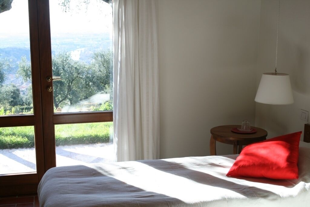 Un dormitorio con cama, almohada roja, mesita de noche con lámpara y vista a la vegetación y un sendero afuera.