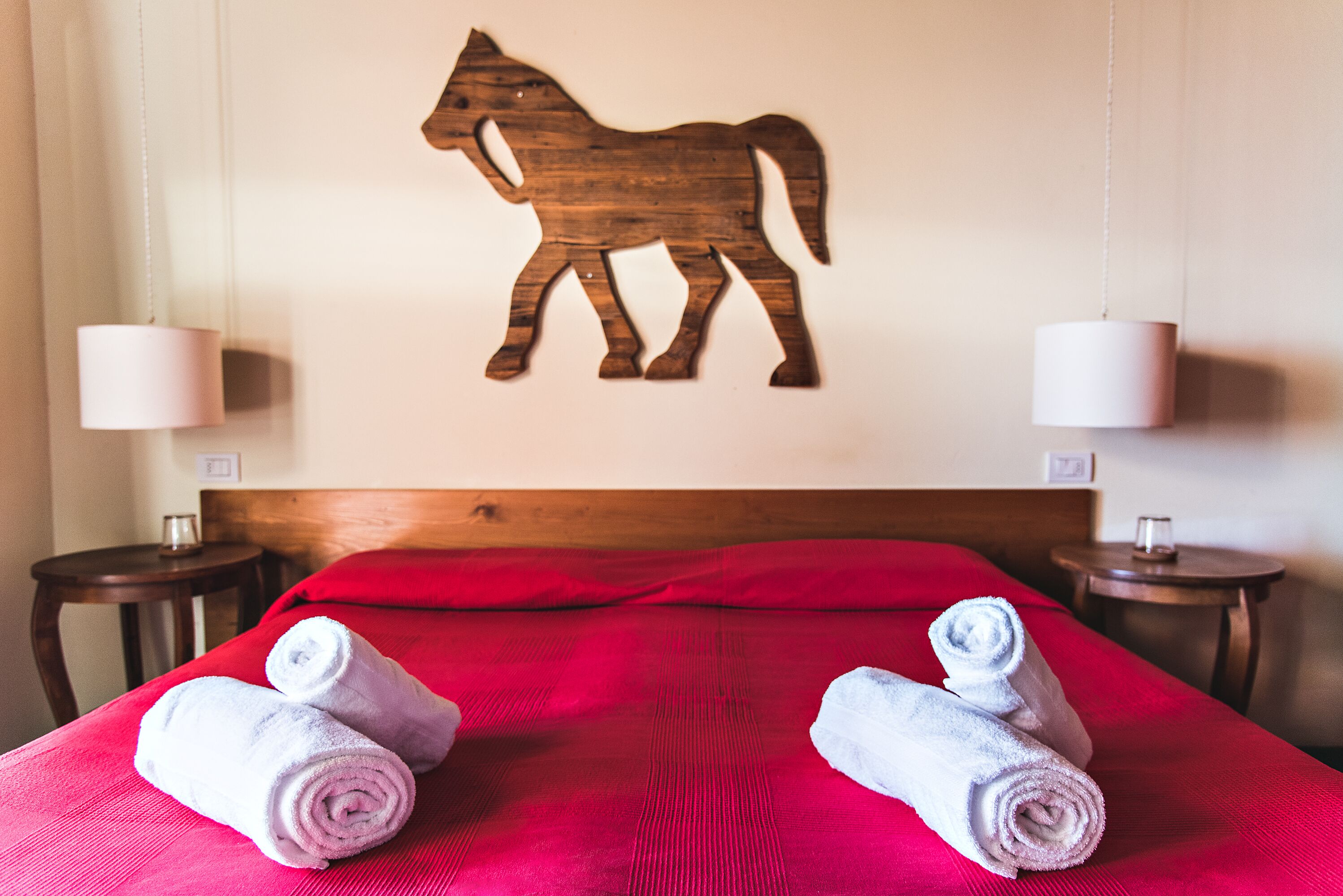 A hotel room with a wooden horse wall decoration, a red bedspread, and two bedside tables with lamps.