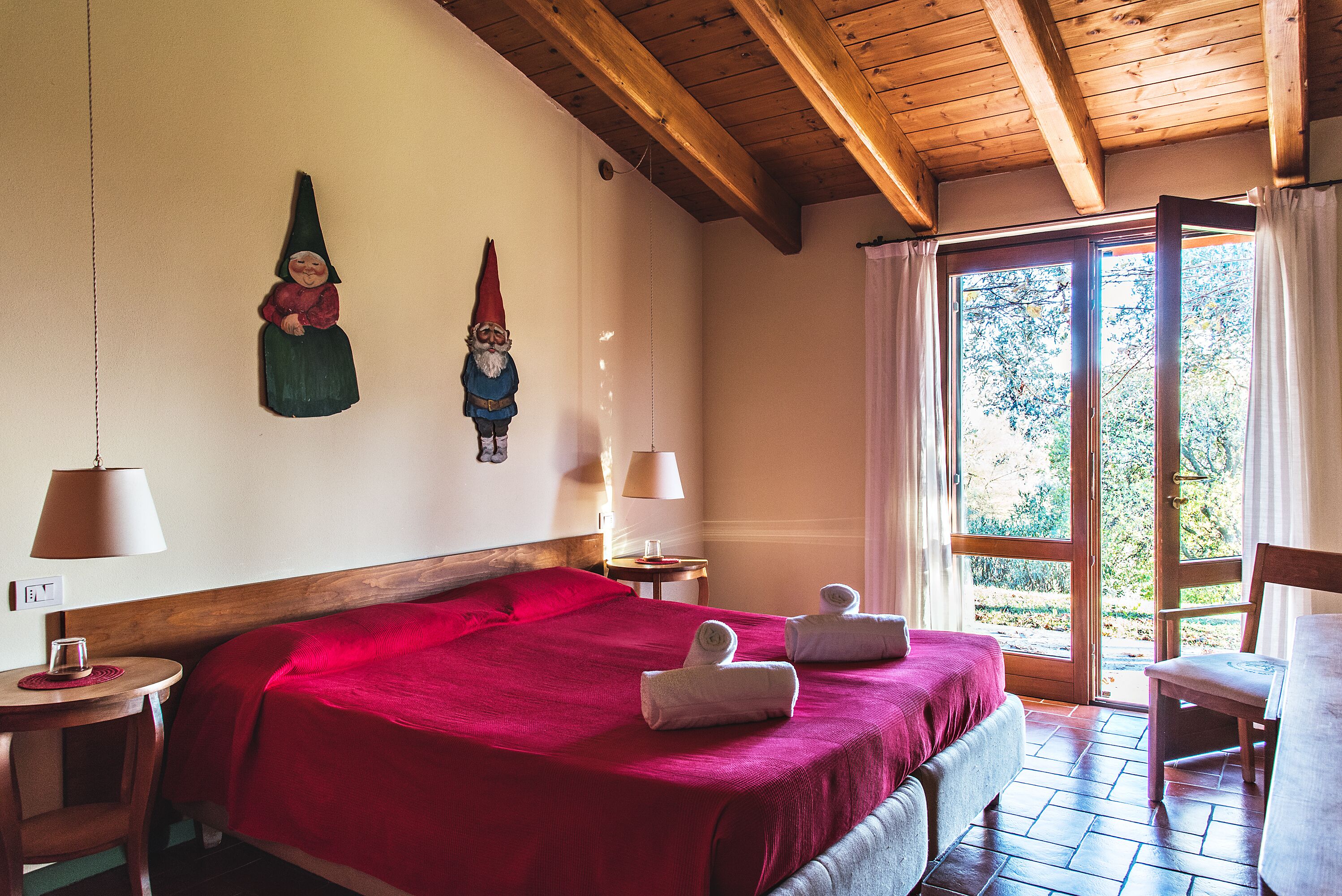 A hotel room with a wooden ceiling, a large bed with a red bedspread, two wall-mounted gnome decorations, a window with curtains, and a small table with a lamp and a glass.