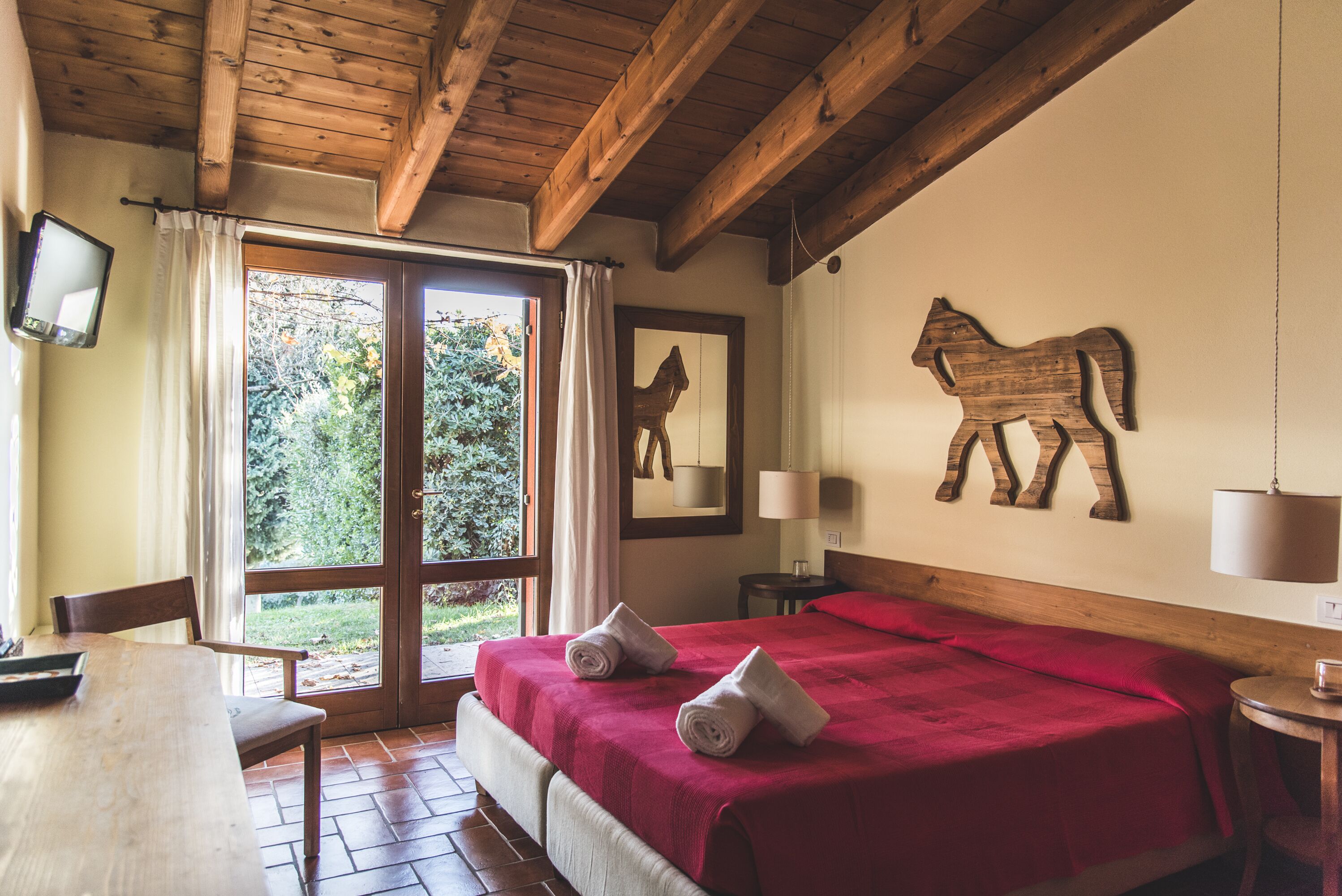 A room with a wooden ceiling, a bed with red bedding, a wooden horse decoration, and a window with curtains.