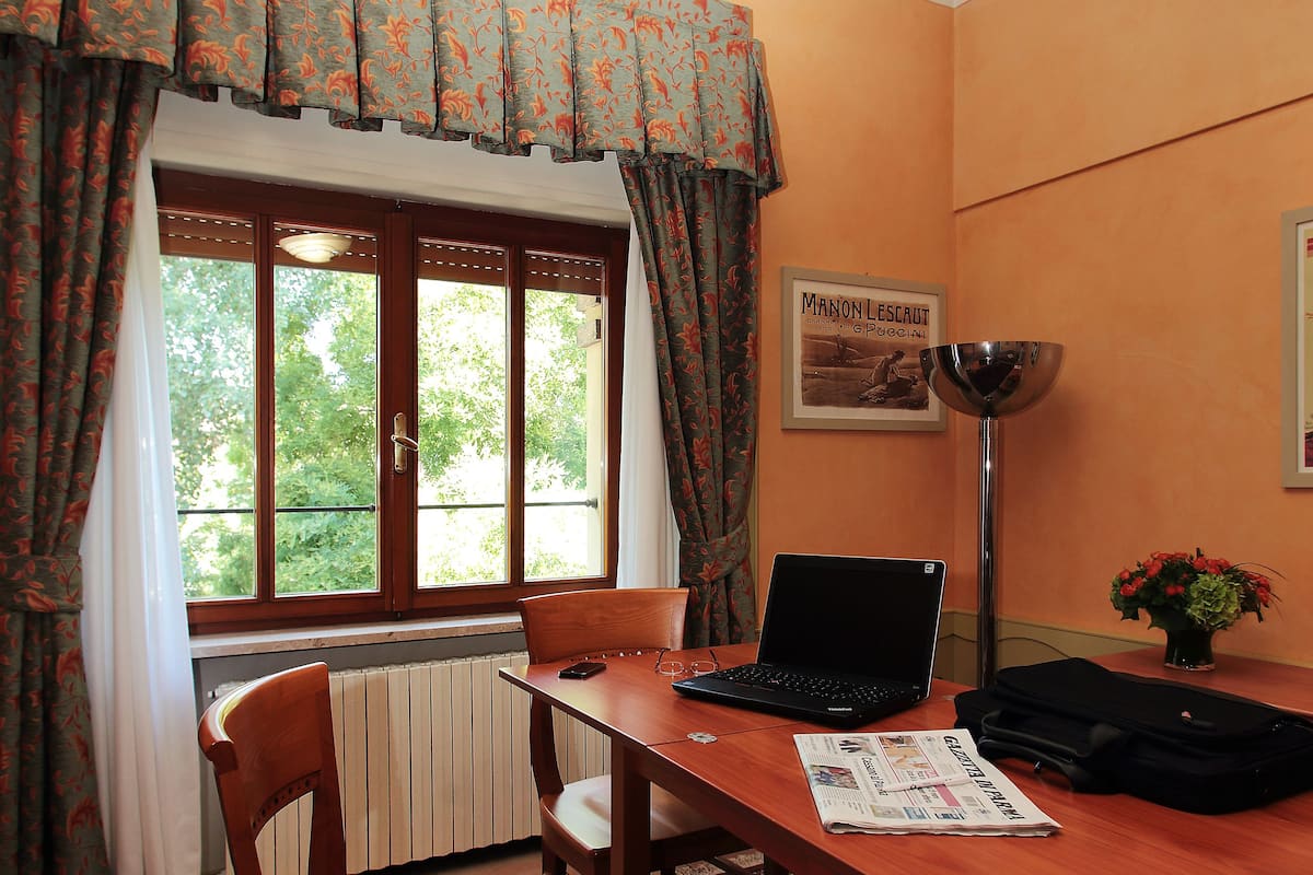 Una habitación con una mesa de madera, una computadora portátil, una silla, un radiador y un ventanal con cortinas.