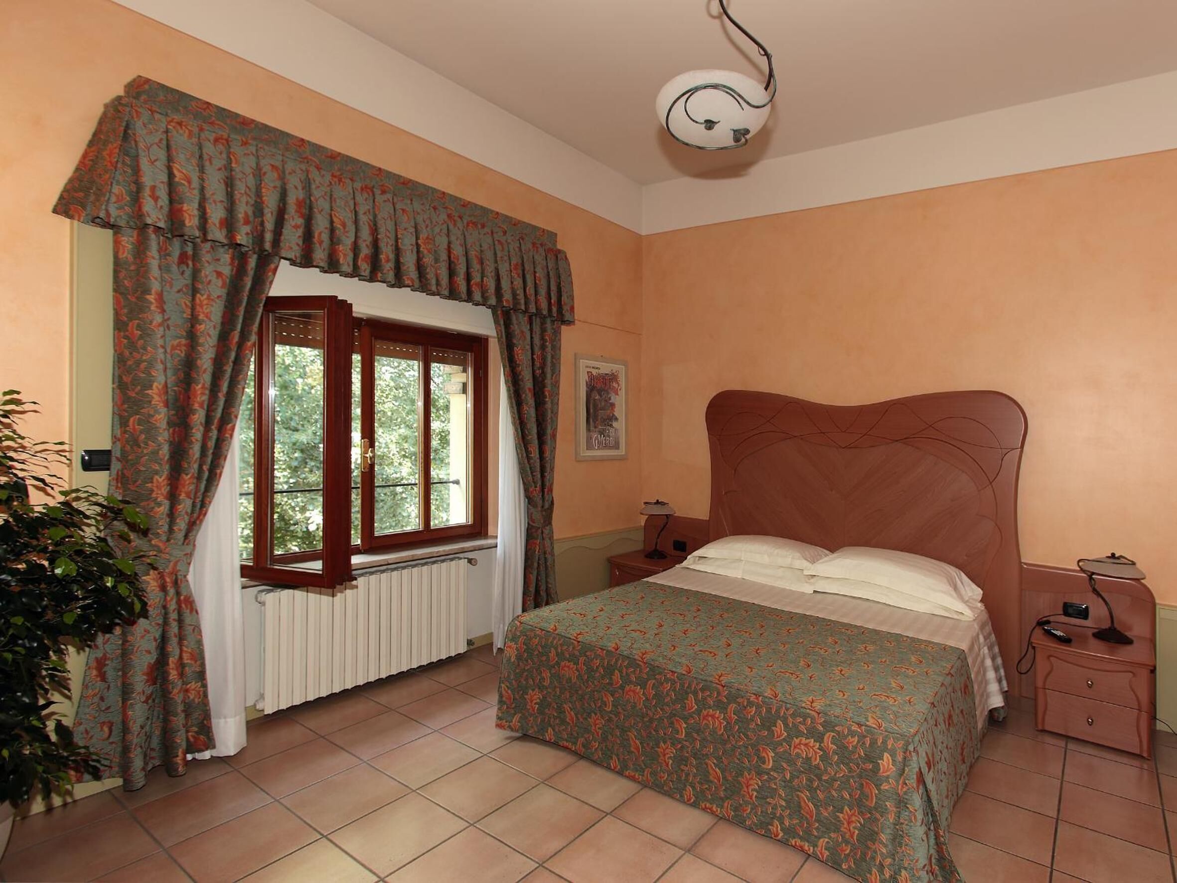 Ein Schlafzimmer mit einem Bett, einem Nachttisch, einer Lampe, einem Heizkörper, einem Fenster mit Vorhängen und einer Deckenleuchte.